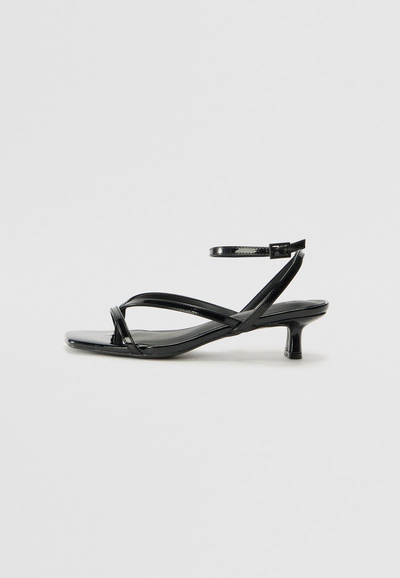 Sandale en cuir verni noir avec un talon bas, présentant des lanières croisées et une sangle de cheville ajustable. Surface lisse et brillante.