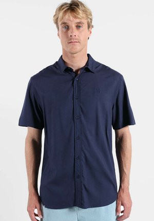 Homme blond aux yeux bleus portant une chemise à manches courtes boutonnée bleu marine et un pantalon bleu clair, debout sur un fond blanc.