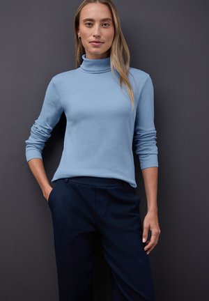 Helles blaues, langärmliges Rollkragenoberteil in ribbeliger Textur, kombiniert mit dunkelblauen, taillierten Hosen. Der neutrale Hintergrund hebt das Outfit hervor.