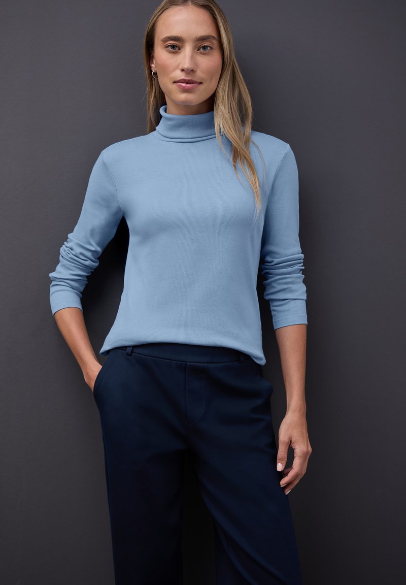 Jasnoniebieski, długi rękaw, golf w ribbowym wzorze, zestawiony z ciemnogranatowymi spodniami o kroju dopasowanym. Neutralne tło podkreśla stylizację.