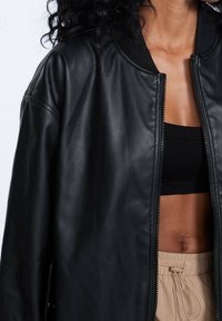 Veste aviateur en cuir noir avec fermeture éclair, col côtelé et coupe décontractée. Portée sur un top court noir et un pantalon beige avec cordon de serrage.