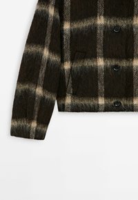 Manche et bas avant d’une veste à carreaux marron foncé duveteuse avec motif à carreaux beige et gris et boutons noirs sur fond blanc.