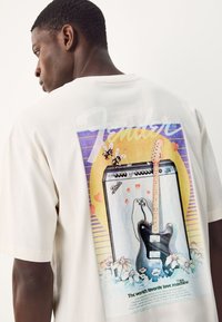 Off-white cotton t-shirt featuring a colorful graphic of an amplifier and guitar, with playful cartoon elements, text, and floral accents.