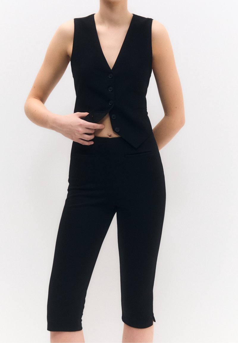 Gilet nero con scollatura a V profonda e sei bottoni, abbinato a pantaloni capri neri a vita alta. Vestibilità aderente, texture liscia con tasche laterali.