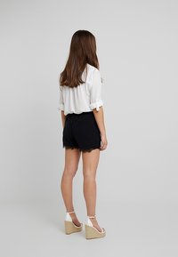 Svarta shorts med spetsdetaljer, tillsammans med en vit skjorta med knappar. Modellen bär beigefärgade kilklackar. Bakifrån mot en enkel bakgrund.