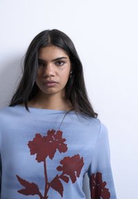 Femme aux longs cheveux bruns portant un haut bleu clair avec un grand imprimé floral rouge foncé, debout contre un mur blanc uni.