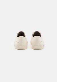 Ett par off-white kanvas-sneakers sedd bakifrån med gummisulor och "ALLSAINTS"-logotyp på hälflikarna.