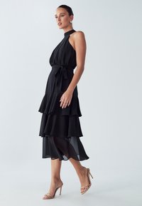 Musta kerroksittain liehuva mekko, jossa on halterneck-kaulus ja vyötäröllä samaa kangasta oleva nauha. Läpinäkyvät kerrokset luovat kerroksellisen efektin; yhdistetty beigenvärisiin korkokenkiin.