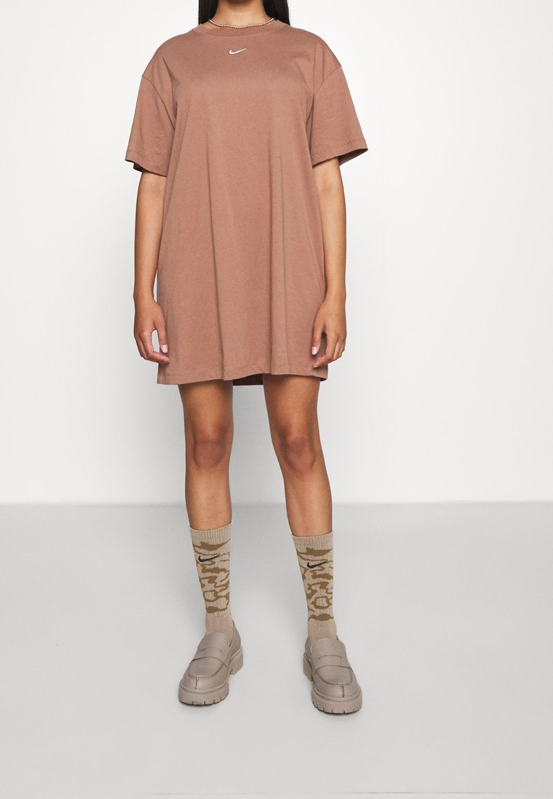 Brązowy, oversize t-shirt z krótkim rękawem, z białym logo Nike. Beżowe skarpetki w wzory oraz masywne beżowe buty z gumową podeszwą.