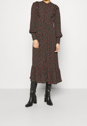 Vrouw draagt een zwarte midi-jurk met lange mouwen, kleine rode bloemenprint en een gerimpelde zoom, gecombineerd met zwarte kniehoge laarzen met hak.