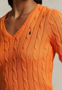 Pull orange en maille torsadée avec un col en V. Présente un petit logo bleu marine sur la partie gauche de la poitrine. Tissu texturé et léger.