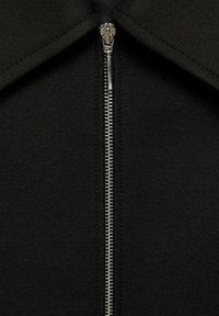 Sort jakke med en central metallynlås, spidse kraver og tekstureret stof, der viser en glat overflade og robuste metaldele.