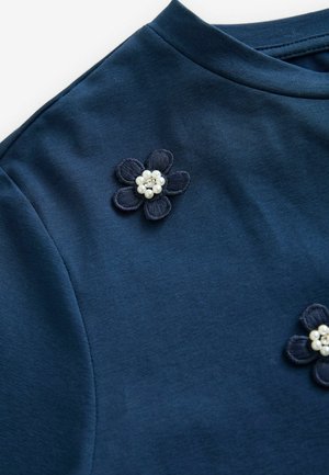 Granatowa bawełniana bluzka z okrągłym dekoltem, zdobiona haftowanymi kwiatami w ciemniejszym odcieniu niebieskiego oraz białymi perłowymi akcentami na lewym ramieniu.