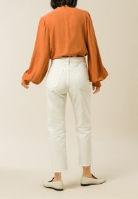 Orangenbluse mit Puffärmeln, kombiniert mit weißen geraden Jeans mit fransigen Säumen und beigen Slippern.