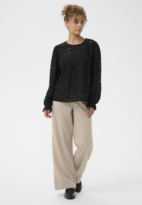 Göndör hajú nő fekete csipkebetétes, hosszú ujjú blúzban, bézs széles szárú nadrágban és fekete cipőben áll egy sima fehér háttér előtt.