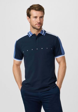 Homme avec une barbe courte et des cheveux bruns portant un polo bleu marine avec des accents bleu clair et le texte "Hackett", debout les mains dans les poches.