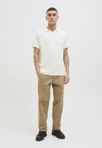 Trumpomis rankovėmis, baltas polo marškinėliai su apykakle ir tekstūruota medžiaga, derinami su smėlio spalvos kelnėmis ir tamsiai rudos spalvos batais, pateikti vienodo fono fone.