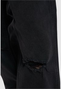 Tejido de mezclilla negra con un desgarro desgastado en el codo, con bordes deshilachados. El material es resistente y tiene una textura suave.