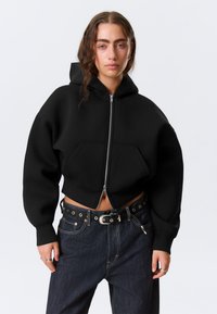 Μαύρο κοντό hoodie με φερμουάρ μπροστά, μεγάλο μπροστινό τσέπη και φαρδιά μανίκια. Το ύφασμα φαίνεται μαλακό και με υφή. Συνδυασμένο με σκούρο τζιν παντελόνι.