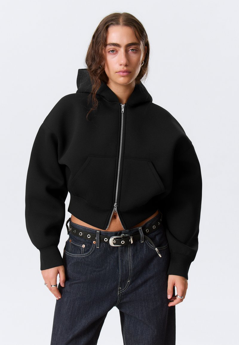 Μαύρο κοντό hoodie με φερμουάρ μπροστά, μεγάλο μπροστινό τσέπη και φαρδιά μανίκια. Το ύφασμ α φαίνεται μαλακό και με υφή. Συνδυασμένο με σκούρο τζιν παντελόνι.