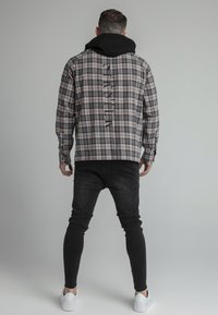 Camicia a quadri con cappuccio nero, caratterizzata da una vestibilità rilassata, schema di colori beige e blu navy e dettaglio di testo verticale sulla schiena. Indossata con jeans neri.