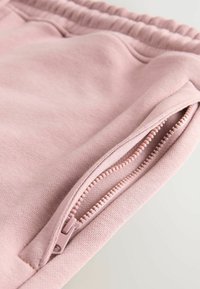 Tejido de sudadera rosa claro con un bolsillo lateral que presenta una cremallera en contraste, mostrando una textura suave y una cinturilla elástica.