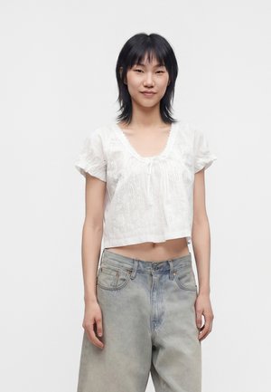 Jeune femme aux cheveux courts noirs porte un chemisier court blanc à manches courtes avec motif œillet et un jean ample en denim clair.