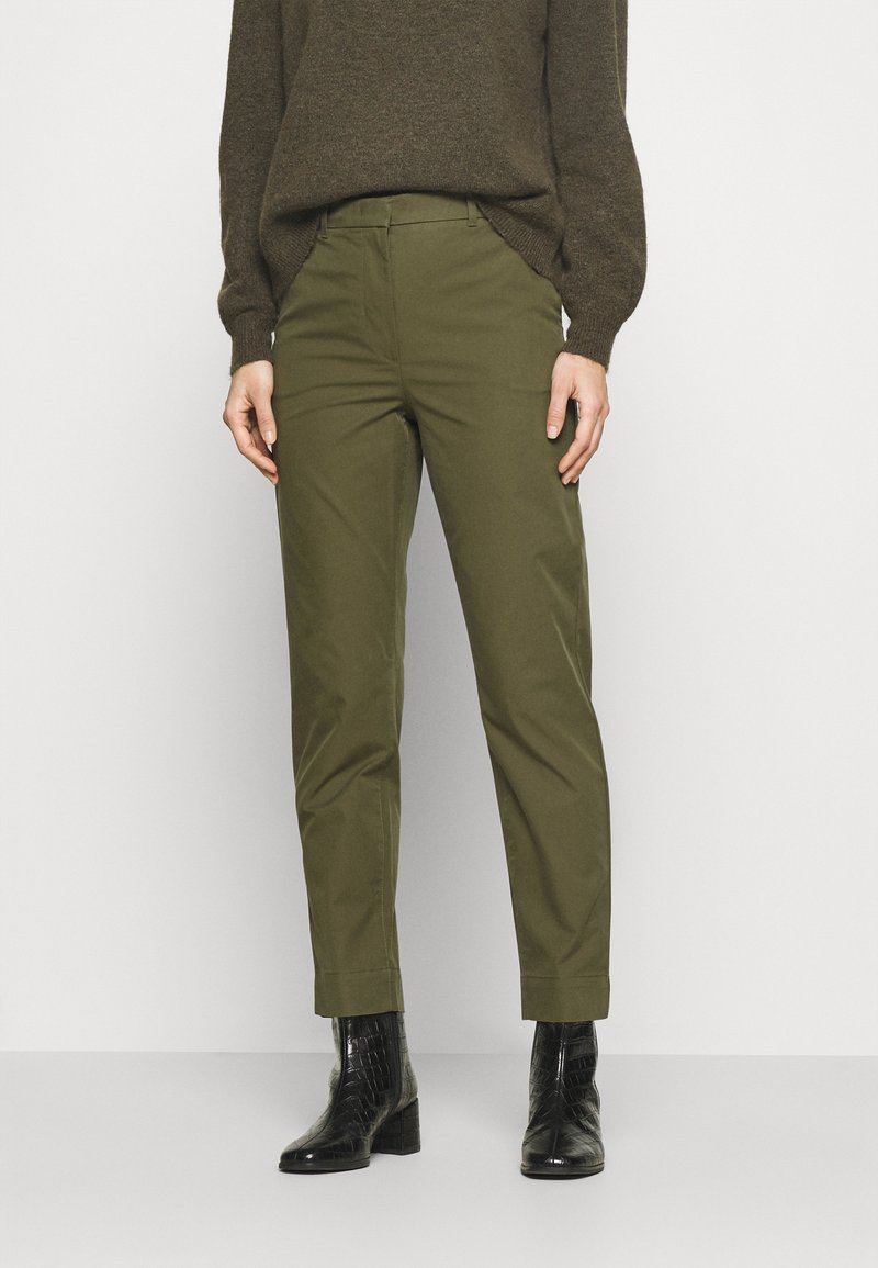 Personne portant un pantalon droit vert olive, un pull marron foncé et des bottines noires à talons sur un fond clair uni.