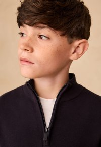 Tumesinine lukuga kampsun kõrge kraega, kootud tekstuuriga ja musta lukuosaga. Allpool on nähtav valge särk. Plekiline nahk.