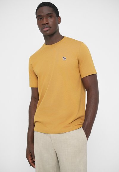 Horčicovo žlté tričko s krátkym rukávom, ktoré má na hrudi malú vyšívanú logo. Materiál vyzerá mäkko a hladko.