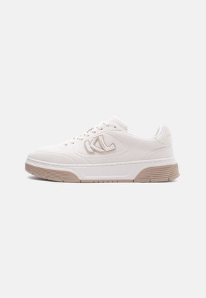 Hvit lave sneakers med beige såle, logo på siden og snøring, på ensfarget lys grå bakgrunn.