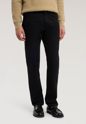 Uomo che indossa pantaloni neri a gamba dritta, mocassini neri e un maglione beige lavorato a maglia, in piedi contro uno sfondo semplice.