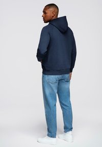 Mężczyzna stoi z rękami w kieszeniach bluzy, ubrany w granatową bluzę, jasnoniebieskie dżinsy i białe trampki na jednolitym tle.