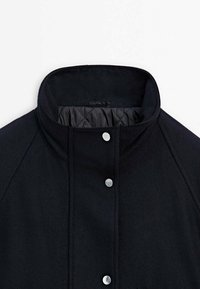 Manteau noir à col montant avec doublure matelassée et trois boutons-pression argentés le long du centre avant.
