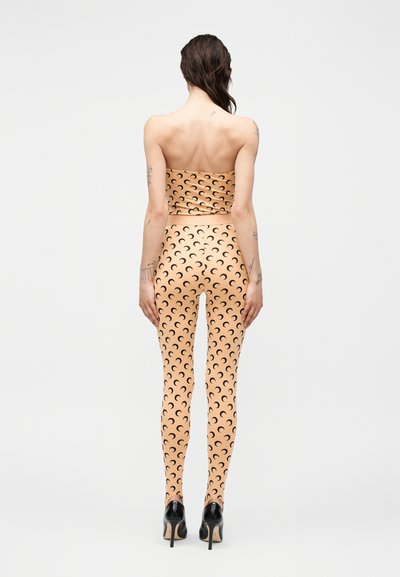 Ženska stoji obrnjena stran, oblečena v tan brezrokavnik in ujemajoče pajkice s črnimi vzorci polmeseca ter črne visoke pete.