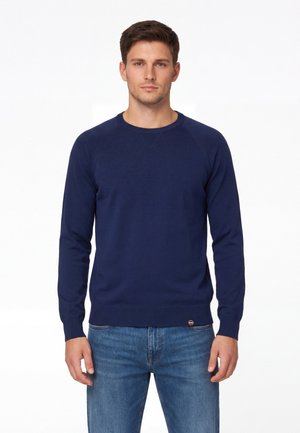 Giovane uomo in piedi, di fronte, che indossa un maglione girocollo blu navy e jeans blu su uno sfondo bianco.