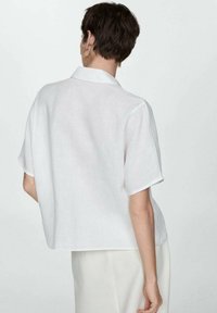 Biała koszula z krótkim rękawem wykonana z lekkiego materiału, z kołnierzykiem i luźnym krojem, o prostych liniach bez widocznych wzorów.