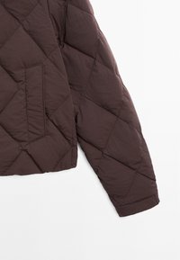 Braune, gesteppte Puffjacke mit einem diagonalen Muster, diagonaler Reißverschlusstasche und weicher Textur. Die Ärmel enden mit einem Bündchen.