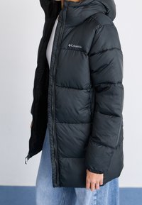 Černá péřová bunda s kapucí, s předním zipem, prošívanou texturou a logem Columbia na levé hrudi. Nositá přes bílou košili.