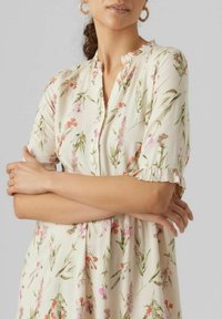 Kwiatowa sukienka w kolorze kremowym z zielonymi i różowymi wzorami, z falbaniastymi rękawami, zapięciem na guziki oraz luźnym krojem. Miękka tkanina o delikatnym połysku.
