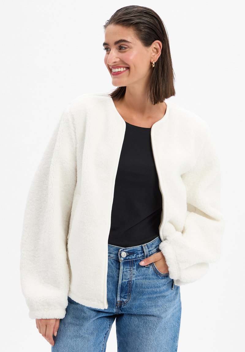 Veste en polaire blanche avec un col rond, fermeture éclair, manches oversized et tissu texturé. Portée par-dessus un haut noir et un jean bleu.