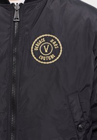 Svart bomberjacka med dragkedja, prydd med en cirkulär guldbroderad logotyp som säger "Versace Jeans Couture". Slät textur.