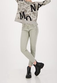 Pull gris clair avec lettres noires, associé à un jean skinny vert clair avec des accents cloutés aux poches et des bottines noires.