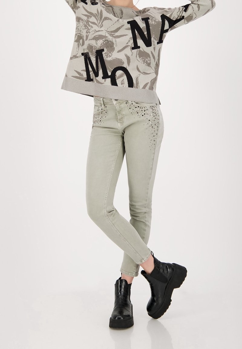 Pull gris clair avec lettres noires, associé à un jean skinny vert clair avec des accents cloutés aux poches et des bottines noires.