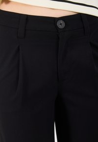 Melnas bikses ar gludu tekstūru, ar priekšējiem krokām, vienu pogu aizdari un sānu kabatām. Pielāgota sagriezuma.