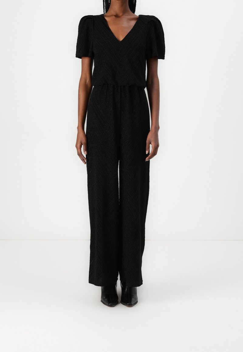 Femme portant une combinaison noire texturée avec des manches courtes bouffantes, un col en V, une taille cintrée, un pantalon à jambes larges et des chaussures noires pointues.