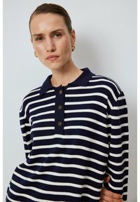 Marinblå och vit randig stickad polo med avslappnad passform, tre-knapps placket och ribbad krage och manschetter. Texturerad tygdetalj synlig.