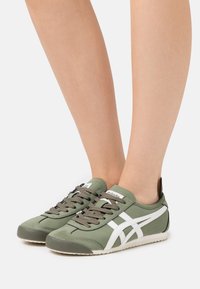 Zapatillas deportivas verdes con un acabado suave, franjas laterales blancas y cordones oscuros. La suela es texturada para mejorar la tracción.