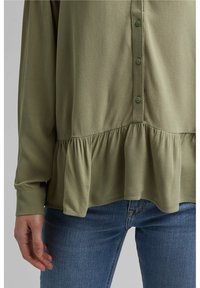 Blusa verde oliva in tessuto leggero, con bottoni frontali e orlo arricciato, maniche lunghe e una vestibilità rilassata sopra jeans di denim.