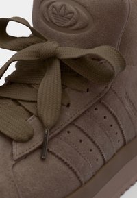 Brune semskede højt-top sneakers med tekstureret overflade, rundt logo, tykke snørebånd og tre stribede accenter på siden. Flad gummisål.